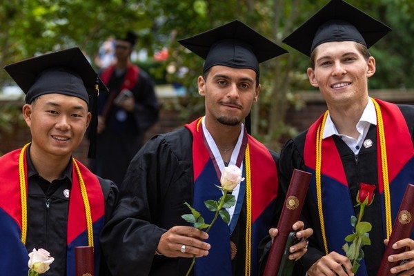 Three graduates in caps and gowns smile outdoors while holding diplomas, standing together with trees and campus buildings in the background.