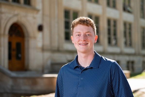 Sam Vinson wears a blue shirt and stands in front of the Engineering building on the University of Arkansas campus.