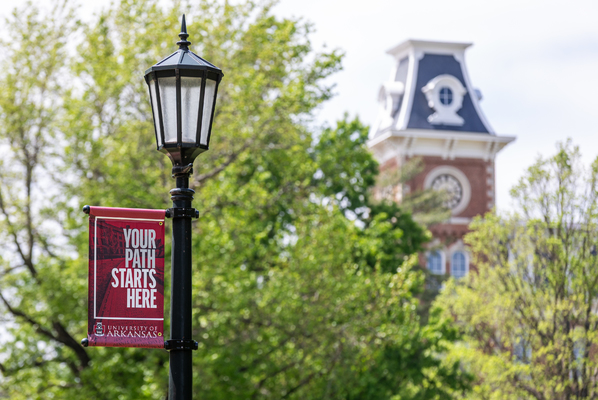 A red banner that says "Your Path Starts Here" is attached to a lamp post in front of Old Main on the University of Arkansas campus. 