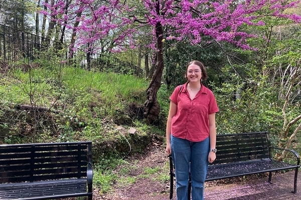Jane Tucker wears a red shirt and blue jeans and stands in front of a park bench. A tree with purple blooms is in the background. 
