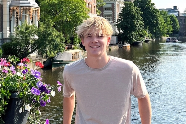 Joseph Hill has blonde hair and a gray tshirt and stands in front of a river with flowers and houses in the background. 