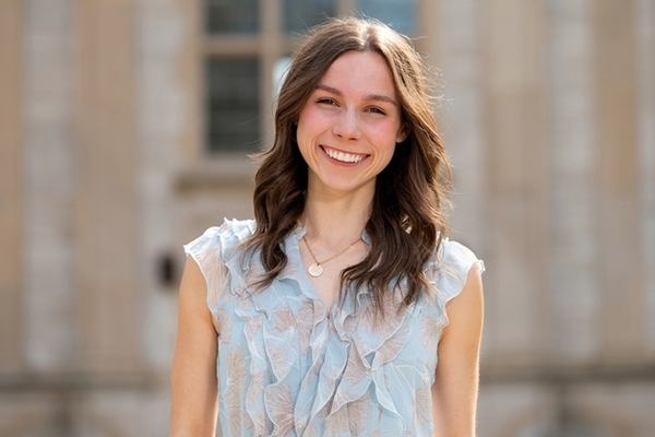 Student stands in front of gray building, smiling, wearing a pale blue sleeveless shirt and has shoulder length brown hair.