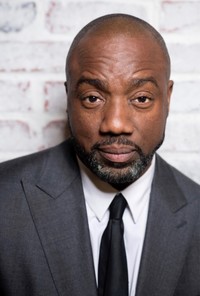  A man in a gray suit and black tie stands against a white brick wall