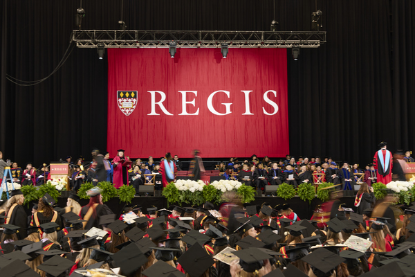 Graduates sit in the audience in front of the Commencement stage that has a large Regis banner.
