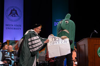 Fighting Okra mascot with large sheet of paper reading "Okra Speech" being assisted by President Dr. Dan Ennis.