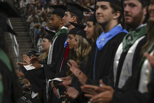 Photo of multiple students in graduation robes standing by their seats at commencement