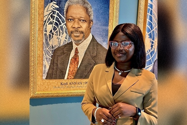 student is wearing tan suit standing in front of portrait with large gold frame. 
