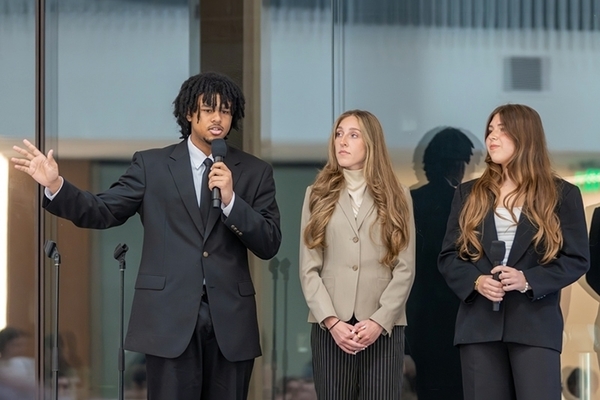 Three students stand in front of a glass panel presenting to crowd. One student is holding a microphone. 