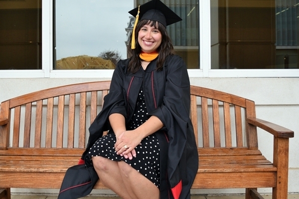 Student sits on wooden bench in her cap and gown. 