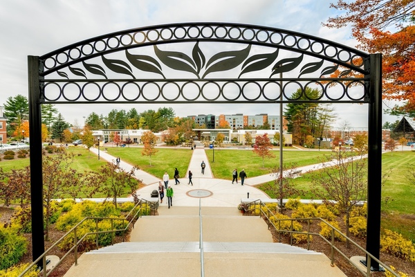 View of the SNHU campus framed by a decorative archway, with intersecting walkways, green lawns, and autumn trees as students walk in the background.