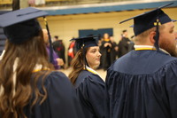 Three graduates in cap and gown stand in processional line. Two females with long brown hair and one male.