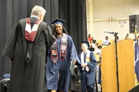 A woman with a decroative stole walks across the stage and shakes hands with a male person wearing a master's degree robe and stole.