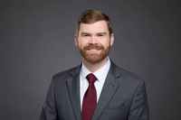 Professional headshot of a man in a suit and tie in front of a gray backdrop.