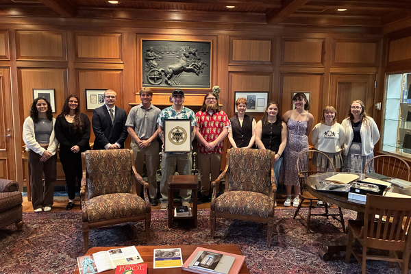 Students stand together in a formal room with wooden panels and antique furniture. A male student in the middle holds a framed Pi Alpha Theta logo. 