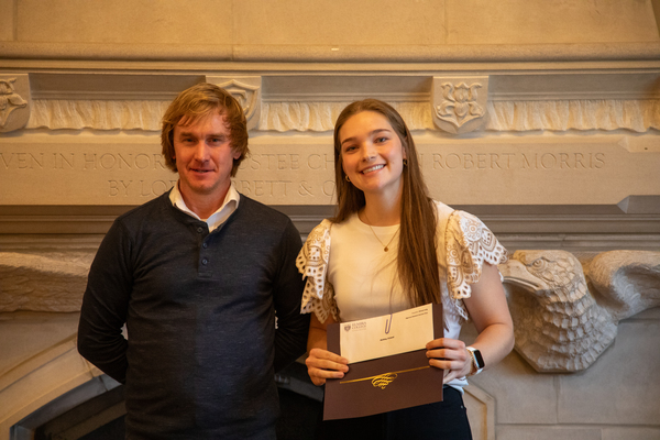 A man dressed in a polo and sweater stands with a young woman with long hair, a creamy white top with lacey sleeves, and a certificate in her hands.