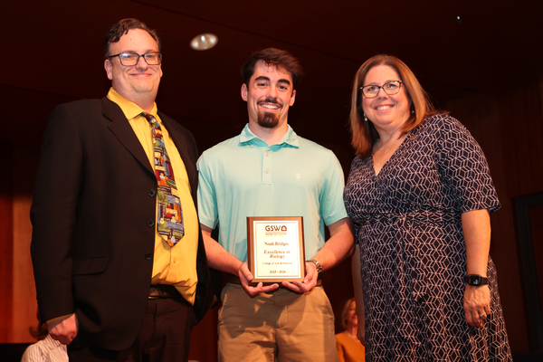 Student poses with plaque in between two faculty members