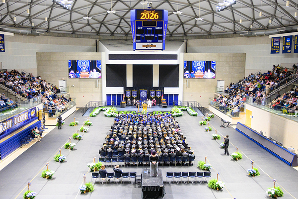 Graduates and faculty sit on the floor of a basketball arena with flowers lining floor and supporters in the stands.