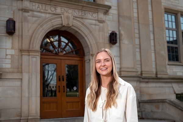 student with blonde hair wears white shirt and stands in front of engineering building on the University of Arkansas campus