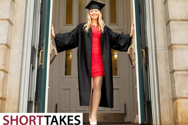 Student stands in red dress and black cap and gown in front of building on the University of Arkansas campus