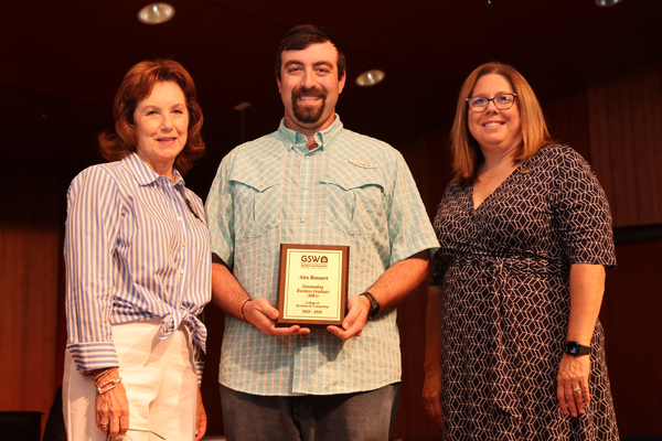Student poses with plaque in between two faculty members