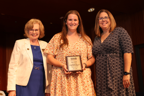 Student poses with plaque in between two faculty members