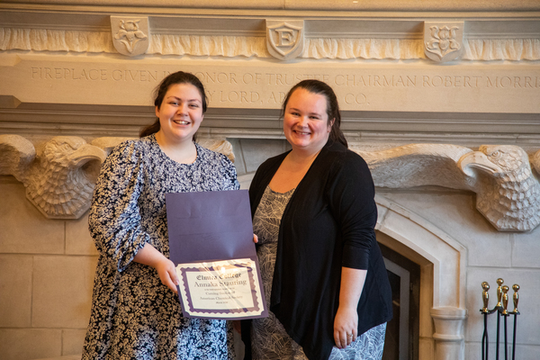 A young woman with long brown hair and a flower dress holds a certificate. Next to her is another woman with brown hair, a dress, and black cardigan.