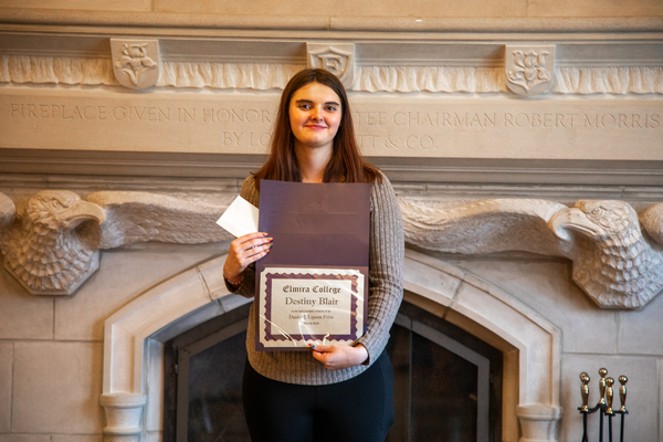 A young woman with long brown hair and a grey top holds a certificate with her name on it in front of her. 