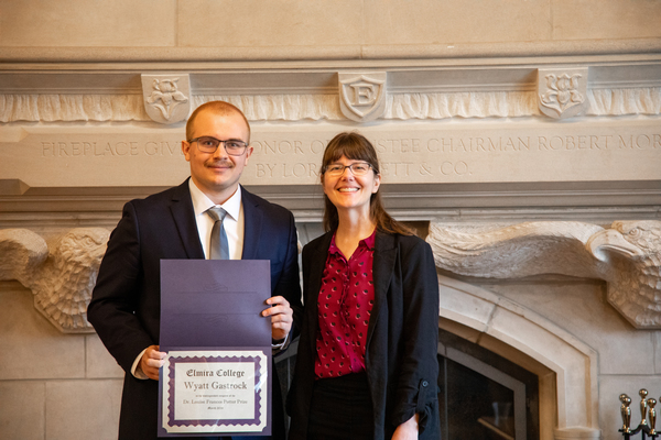 A young man with a shaved head in a suit holds a certificate as he stands next to a woman in a red top and black cardigan. 