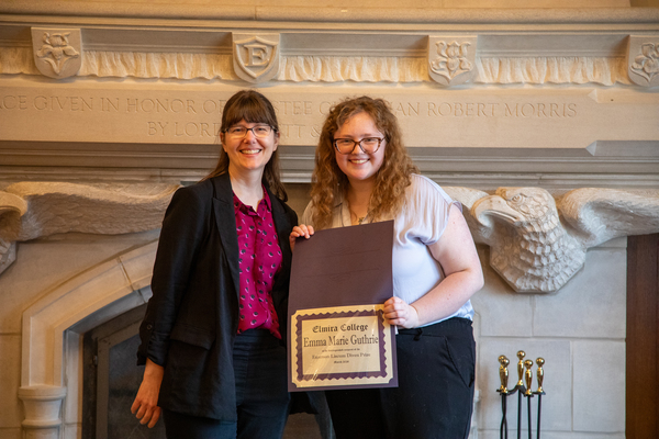 A woman with a red top and black cardigan stands next to a young woman with curly blond hair who is holding a certificate with her name on it. 