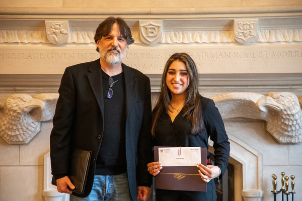 A tall man with brown hair and a white beard in a black shirt and jacket stands next to a woman with brown hair and a black top holding a certificate.