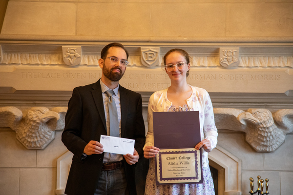 A man with brown hair and beard in a suit stands next to a young woman with light brown hair in a dress who is holding a certificate. 