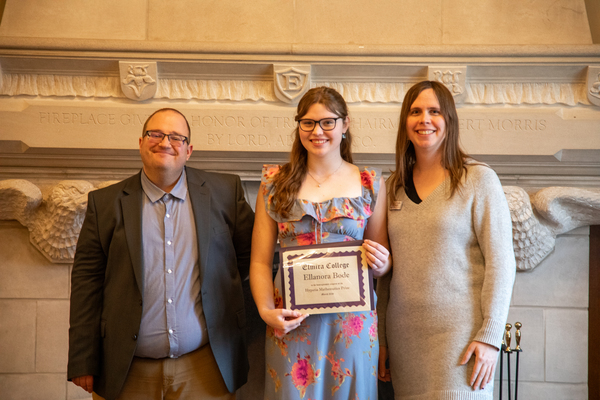 On the left stands a man in a suit. In the middle is a young woman in a dress holding a certificate. On the right is a woman in a light brown dress.