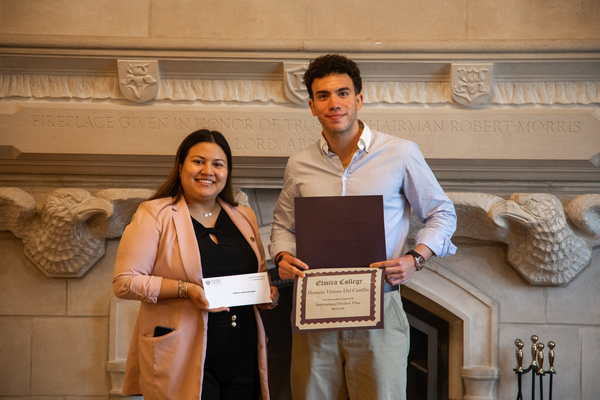 A woman in a long pink coat holds an envelope next to a man with brown hair in a dress shirt who is holding a certificate with his name on it.