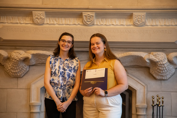 A young woman with brown hair, glasses, and a blue-flowered top stands with a woman with brown hair in a yellow top who is holding a certificate.