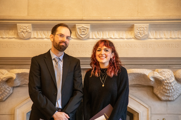 A man with brown hair and a beard in a suit stands next to a young woman with reddish-brown wavy hair in a black top.