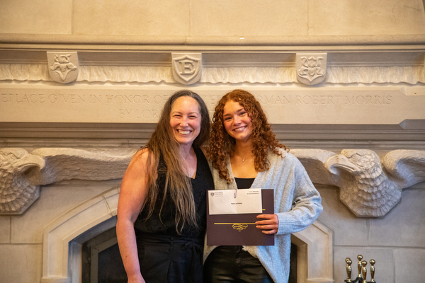 A woman with long light-brown hair in a black dress stands with a young woman with curly red hair in a black top and light-grey sweater. 