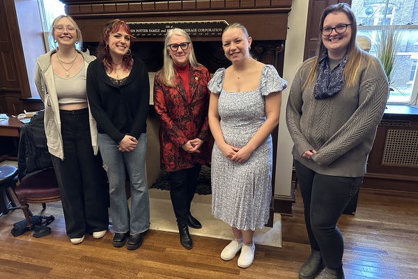 Five women stand shoulder to shoulder, smiling at the camera. They are in front of a fireplace and windows in a room with hardwood floors.  