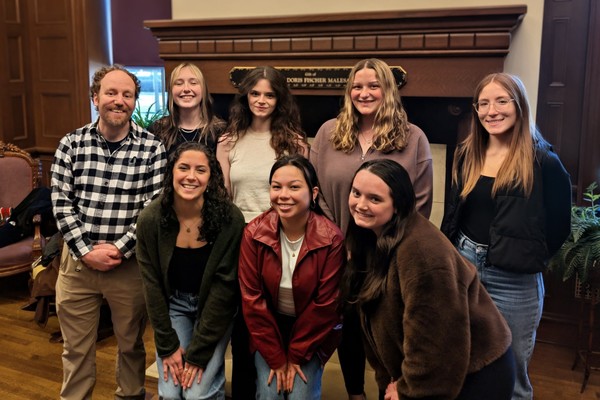 A group of students and a professor stand shoulder to shoulder in two rows as they pose and smile for the camera. 