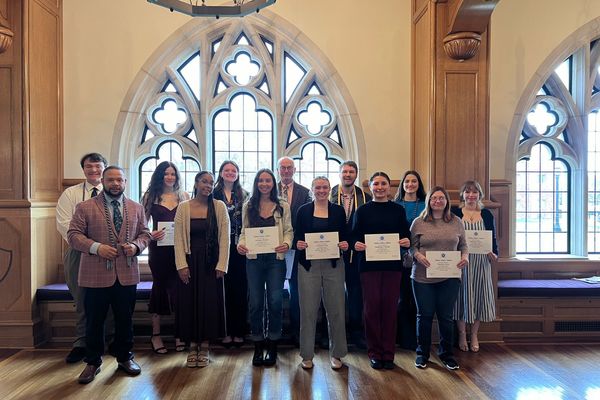 A group of college students stands together in front of a Gothic-style window. The students are all holding certificates in front of themselves. 