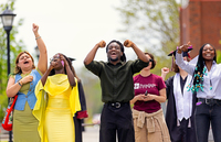 Clad Sanon cheers and pumps his fists in the air at Commencement, standing near other students and guests.