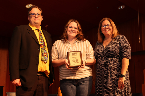 Student poses with plaque in between two faculty members