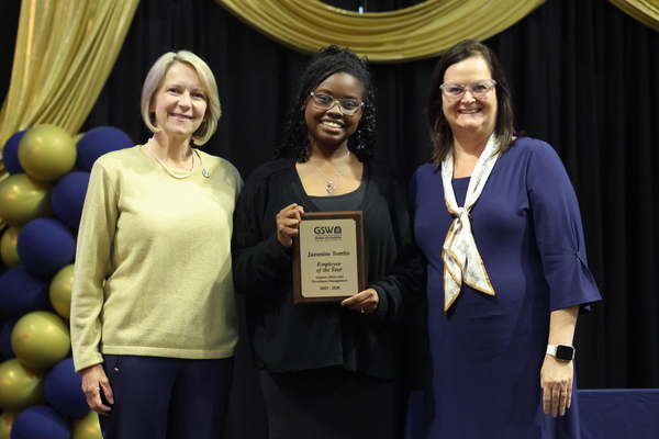 Student poses with plaque in between two staff members