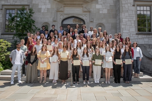 94 students stand in front of gray brick building on the University of Arkansas campus holding certificates. 