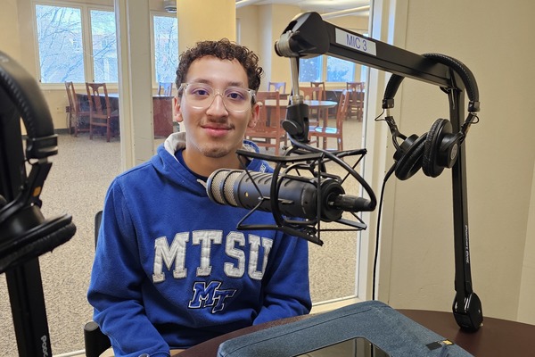 A student wearing a blue MTSU hoodie sits at a podcast recording station with a microphone and headphones visible in the foreground. 