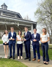 A group of students in business attire pose for a photo in front of the Ives Park gazebo holding a trophy.