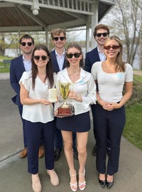 A group of students in business attire and sunglasses pose for a photo in front of the Ives Park gazebo holding a trophy.