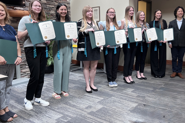 A group of students hold certificates at an indoor ceremony, with a professor on each side of the group.  
