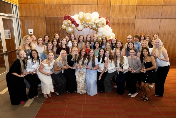 Students stand in a group and pose for a photo in front of a wood wall and decorative balloons. 