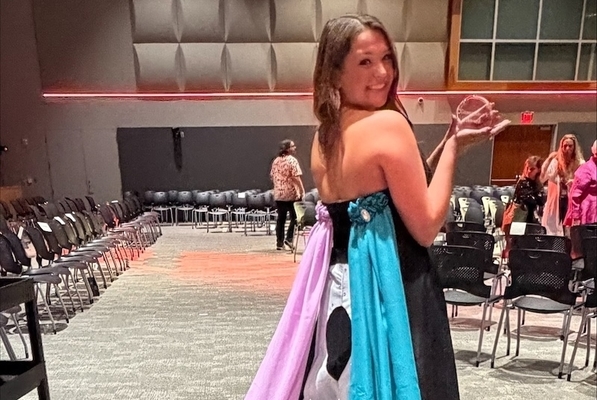 student is waring dress that she designed with pink and blue fabric coming off the back. Student is holding her award at the ceremony. 