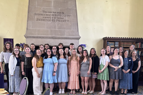 A large group of students dressed nicely stands in front of a large, grey fireplace. The group is looking at the camera and smiling. 
